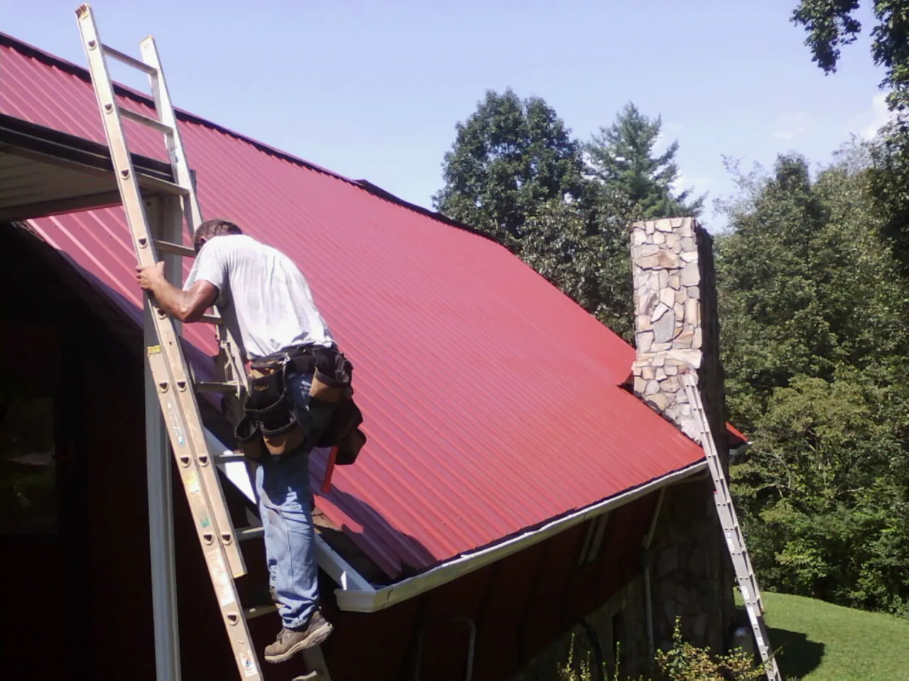 Roofer on a ladder beside a red metal roof and stone chimney — Soffit & Fascia Repair by Roofing Brothers in Scarsdale